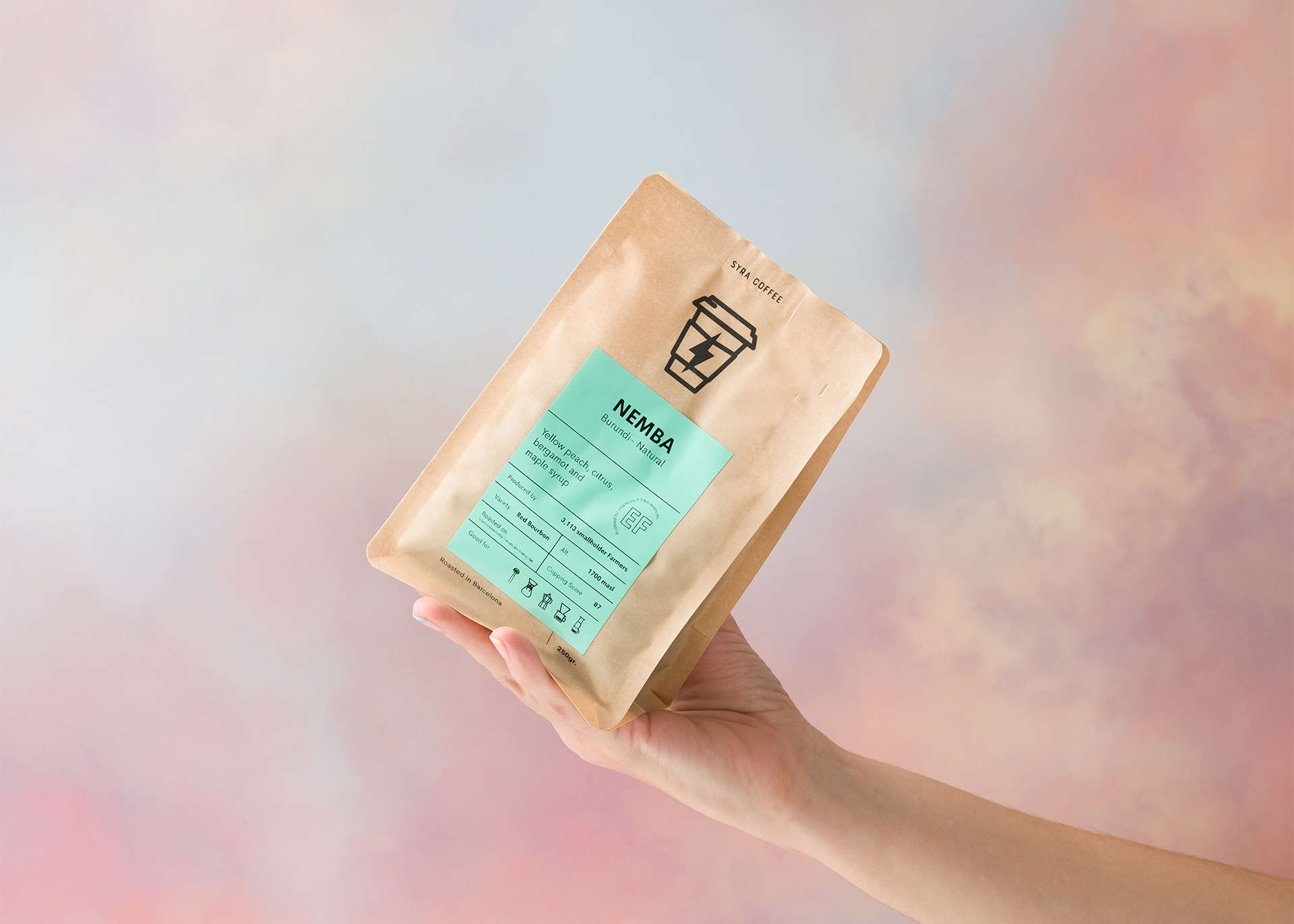 Una mano sostiene una bolsa de café Nemba de Syra Coffee sobre un fondo difuminado azul y rosa